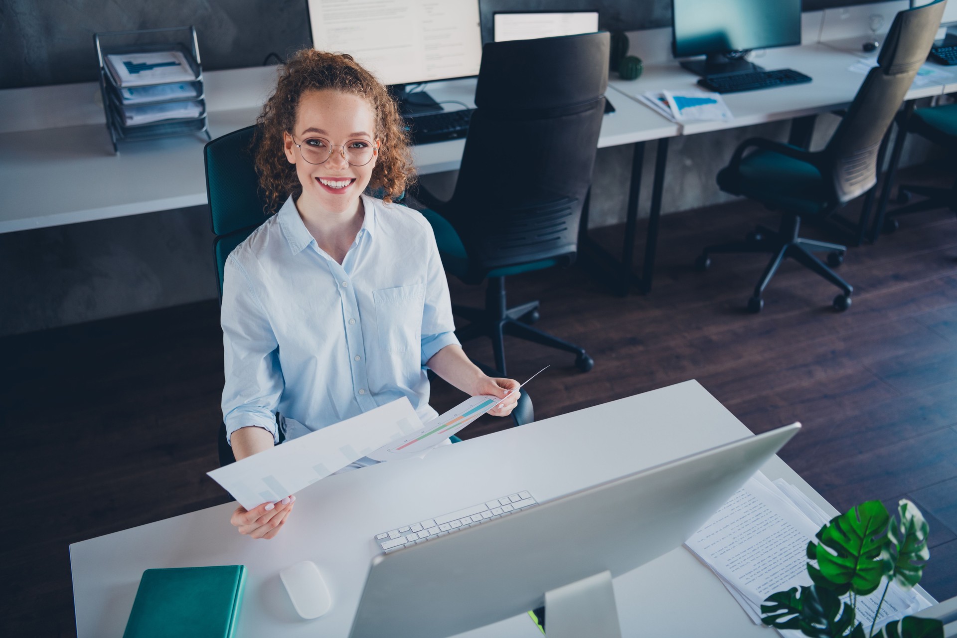 Young businesswoman in formal attire smiling at  workstation, reviewing documents with confidence and professionalism in a modern office